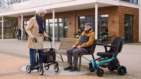 Elektrorollstuhl und Rollator mit zwei Personen an einer Parkbank in einer Fußgängerzone
