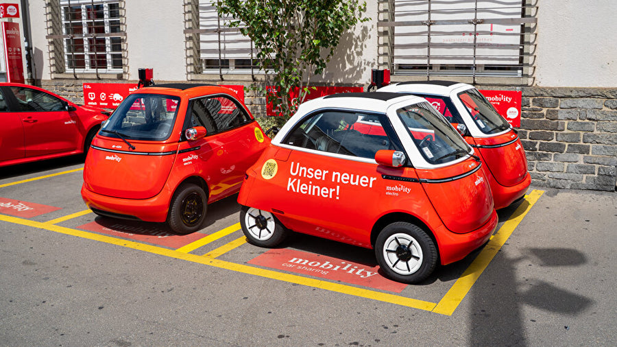 Drei orange-rote Microlino in einer markierten Mobility-Carsharing-Zone in der Stadt